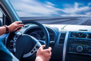 Driver's hands on steering wheel as car speeds down the road under a bright sk