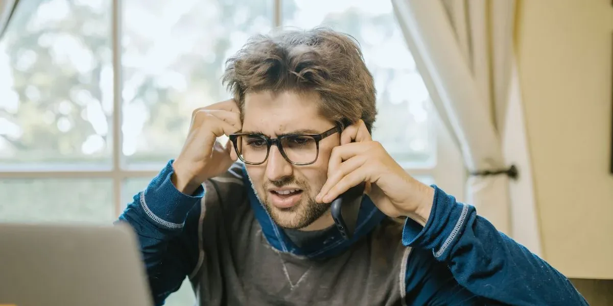 Frustrated Driver on the Phone After Discovering Incorrect Fault Assignment (1) A young man wearing glasses looks frustrated while on the phone and checking his laptop, possibly disputing an auto insurance error.